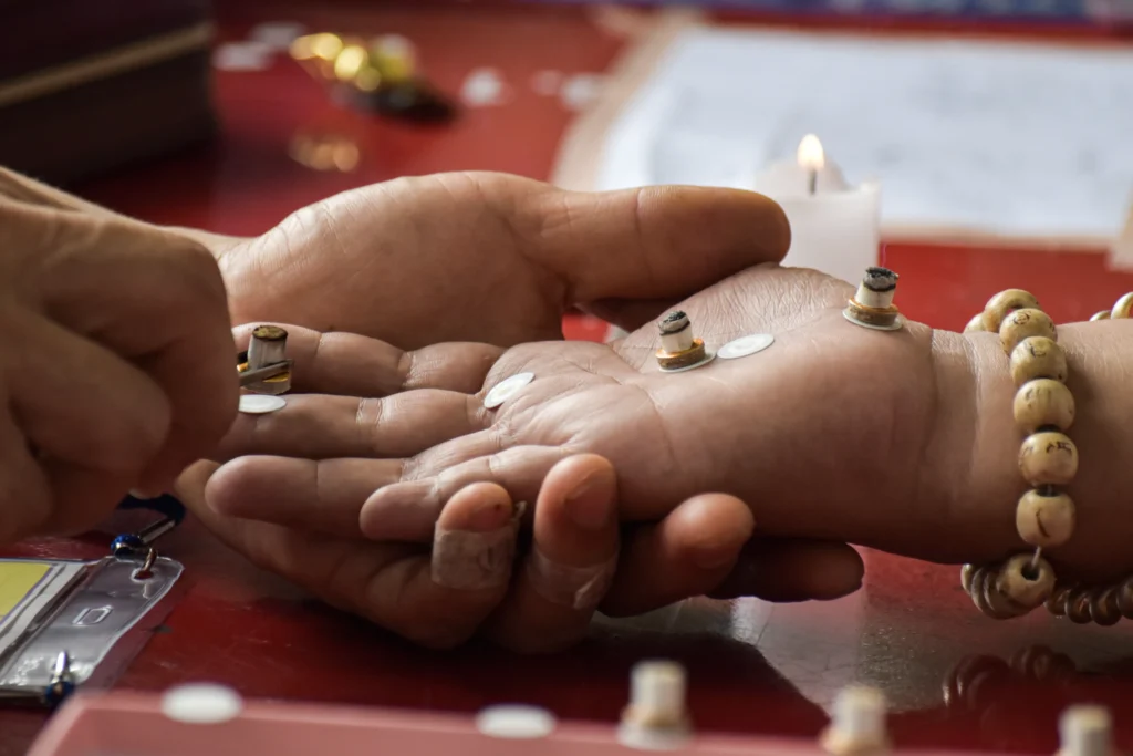 A close-up of a person's palm with small objects and disks placed on it, while another hand interacts; a lit candle and beads are visible in the background.