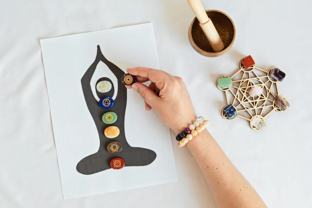 A hand places a crystal on a chakra-themed illustration, surrounded by various gemstones and a singing bowl on a white table.
