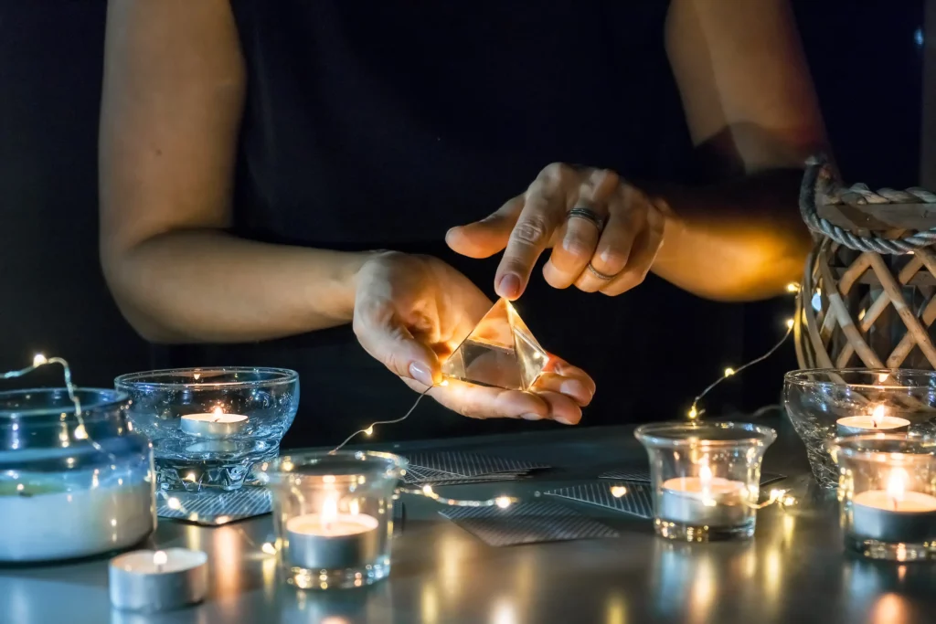 A hand holds a glowing crystal pyramid, surrounded by candles and fairy lights, creating a mystical ambiance.