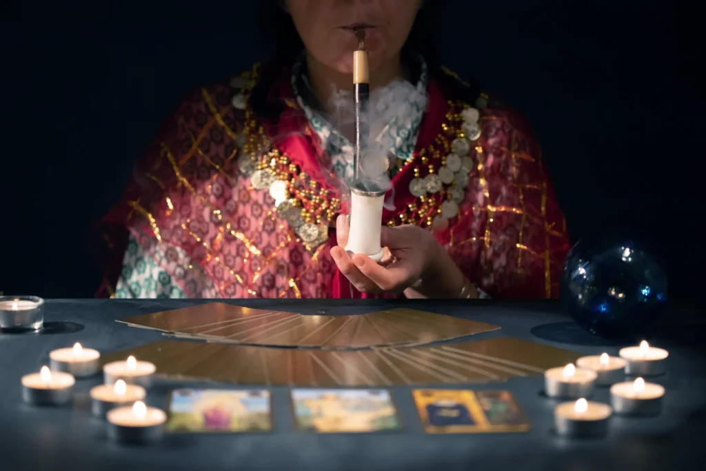 A fortune teller in a vibrant robe blows smoke from a pipe, surrounded by tarot cards, candles, and a crystal ball on the table.